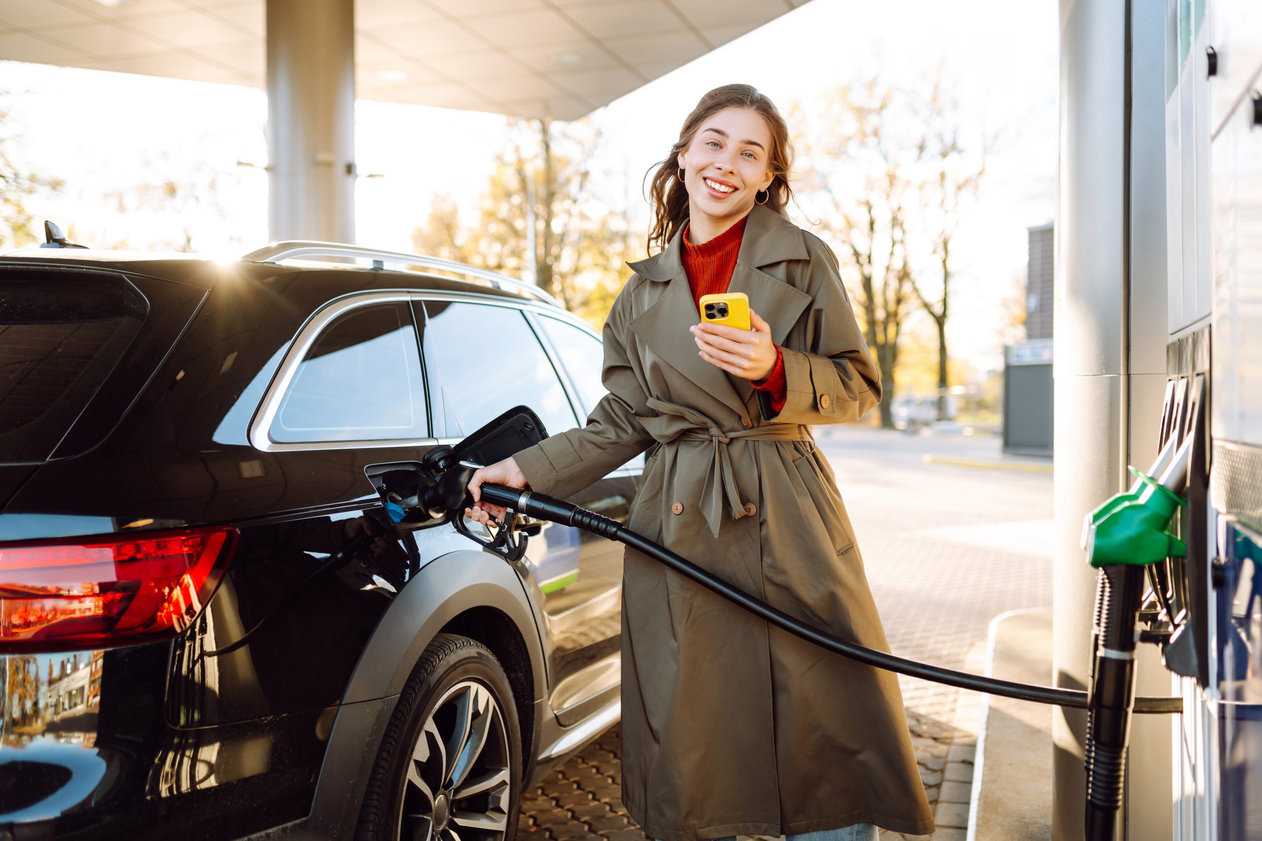A young woman with a phone fills up her car at a gas station. A woman at a gas station fills up her tank with fuel using an app on her phone. Transport concept, industry.