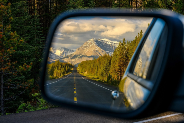 Eine Straße im Jasper-Nationalpark führt durch dichten Wald auf die schneebedeckten Rocky Mountains zu, gesehen im Außenspiegel eines Autos.