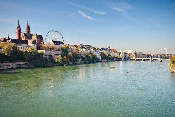Panoramablick auf Basel mit seinen historischen Gebäuden am Rheinufer, einer Brücke über den Fluss und einem Riesenrad im Hintergrund.